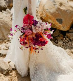 a bride holding a bouquet of flowers in her hand on the rocks by the water