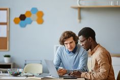 two men sitting at a table looking at a laptop computer and working on something in front of them