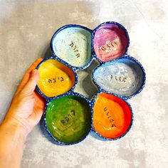 a person holding their hand in front of six bowls with different colors on the surface