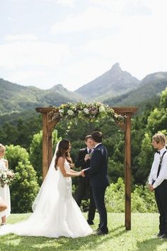 a bride and groom standing in front of a wooden arch with greenery on it