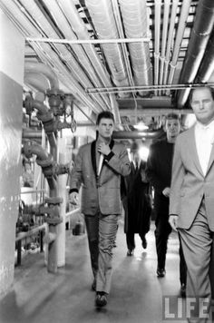 black and white photograph of men in suits walking down a hallway with pipes on the ceiling