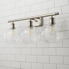 three clear glass globe lights hang from a metal bar on a white tiled wall in a bathroom