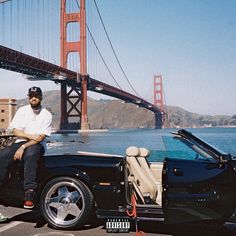 a man sitting on the hood of a black sports car in front of a bridge