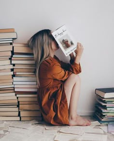 a woman sitting on the floor reading a book with stacks of books behind her, all around her