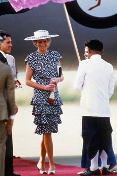 a woman in a dress and hat standing under an umbrella with other people around her