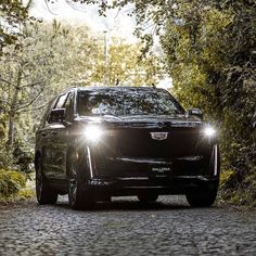 the front end of a black suv driving down a road surrounded by trees and bushes