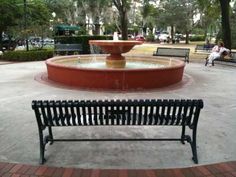 an empty park bench in front of a fountain with people sitting on benches around it