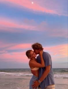 a young man and woman embracing on the beach at sunset, with pink clouds in the background