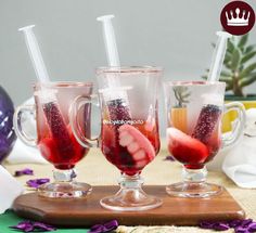 three glasses filled with liquid sitting on top of a wooden cutting board next to purple flowers