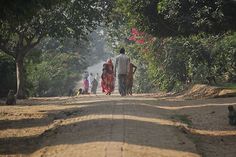 two people walking down a dirt road with trees in the background