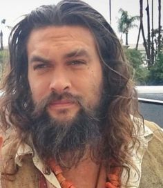 a man with long hair and beard wearing an orange beaded necklace is standing in front of a car