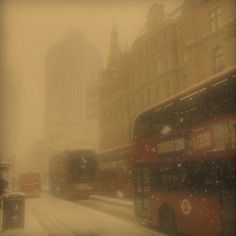 two double decker buses driving down a snow covered street with tall buildings in the background