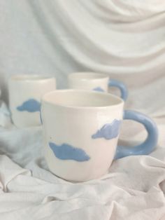 three white and blue coffee mugs sitting on top of a bed covered in sheets