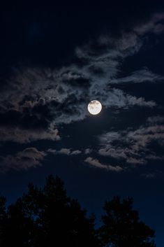 the moon is shining brightly in the night sky above some trees and dark cloudy skies