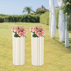 two tall white vases with pink flowers in them on the grass near an archway