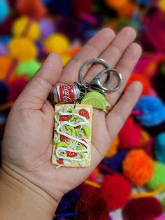 a hand holding a tiny sandwich keychain with a lime slice on it and a soda can in the other hand