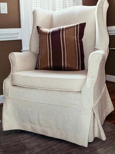 a white chair with two pillows on top of it in front of a window and wooden floor