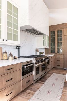 a kitchen with white walls and wooden floors has stainless steel ovens, cabinets, and an area rug on the floor