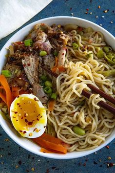 a white bowl filled with noodles, meat and veggies next to an egg