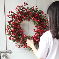 a woman is decorating a wreath with red berries and pine cones on the front door