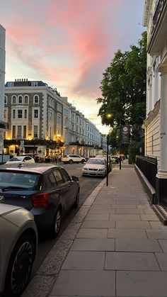 cars parked on the side of a street next to tall buildings and trees at dusk