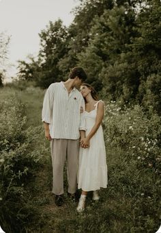 a man and woman standing next to each other in the grass with trees behind them