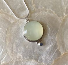 This pendant features a natural, round, Green Serpentine gemstone. This particular gemstone has natural, white, polkadot, inclusions that set it apart from the other gemstones. I knew I had to work with it 🥰 The semi-translucent nature and soft green color inspired me to make a simple but unique setting! I began by using a plain, silver, bezel wire, to secure the stone. Then, I attached a medium-size, plain, round, wire to frame part the stone, altering the silhouette of the stone just a bit. Then, I added a small piece of plain, round wire to fit against the other piece of plain wire. I did this to further alter the silhouette. Next, I added a flattened, sterling silver granule to the bottom of the piece. Lastly, I created a single, "V" bale (5mm), allowing for a variety of chains to be Round Jade Cabochon Necklace, Chalcedony Natural Stone Round Necklaces, Jade Cabochon Round Pendant Necklace, Round Jade Cabochon Pendant Necklace, Chalcedony Necklaces With Natural Stones, Elegant Gemstone Round Disc Necklace, Unique Round Chalcedony Necklace, Handmade Round Chalcedony Necklace, Bezel Wire