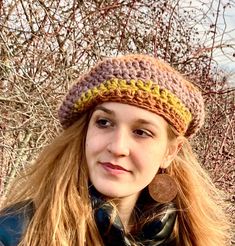 a woman with long hair wearing a crocheted hat and scarf, standing in front of trees