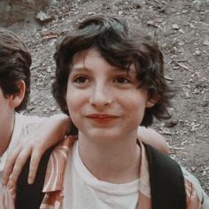 two young boys standing next to each other in front of a dirt ground covered with leaves