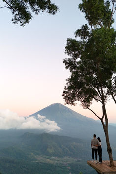 two people are standing on a ledge looking at the mountains and clouds in the distance