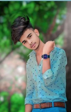 a young man with a blue shirt and brown belt is posing for a photo in front of some trees