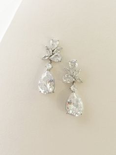 two pairs of diamond earrings on a white surface with water drops in the center and leaves at the bottom