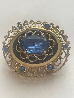 Immerse yourself in vintage elegance with this exquisite signed Coro Pegasus MCM royal blue rhinestone domed brooch. This stunning piece showcases a magnificent royal blue rhinestone center, surrounded by intricate gold-tone filigree and additional blue rhinestones, creating a breathtaking visual effect that embodies mid-century modern sophistication. Imagine this brooch gracing the attire of a stylish individual in the mid-20th century, adding a touch of regal elegance to any outfit. The bold b Blue Victorian Brooch For Formal Occasions, Victorian Blue Brooch For Formal Occasions, Blue Victorian Brooches For Formal Occasions, Vintage Sapphire Brooch Jewelry, Ornate Blue Brooch For Formal Occasions, Blue Oval Brooch, Ornate Blue Brooches For Wedding, Ornate Blue Wedding Brooches, Blue Oval Brooch For Formal Occasions