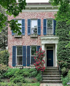 an old brick house with blue shutters and red roses on the front door is surrounded by greenery