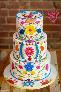 three tiered cake decorated with colorful flowers on a wooden table next to a brick wall