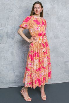 a woman standing in front of a gray wall wearing a pink and orange floral dress