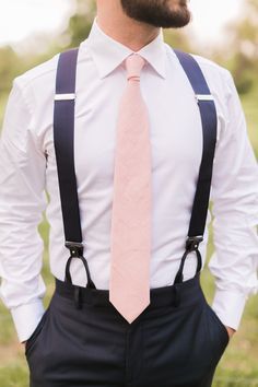 a man wearing suspenders and a pink tie is standing in the grass with his hands on his hips