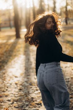 a woman standing in the middle of a leaf covered road with her hair blowing in the wind