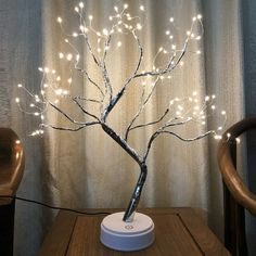 a small tree with white lights is on a table next to a chair and curtain