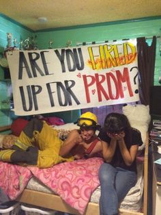 two people sitting on a bed with a sign that says are you fired up for prom?