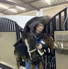 a woman standing next to a brown horse in a stall with another horse behind her