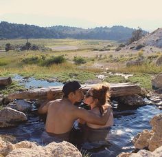 a man and woman are sitting in the water near some rocks, grass and trees