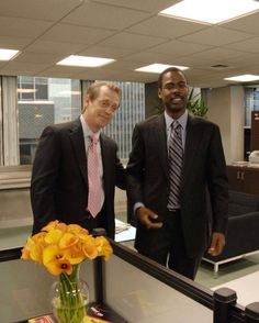 two men in suits and ties standing next to each other near a vase with flowers