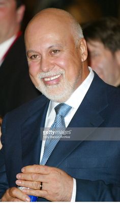 an older man in a suit and tie smiles as he holds his hand on his chest