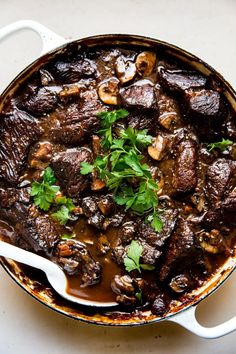 a pot filled with meat and mushrooms on top of a white countertop next to a wooden spoon