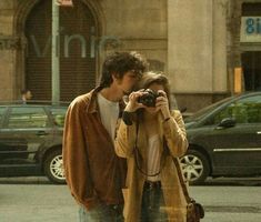 a man and woman walking down the street while taking pictures with their cell phones in front of them