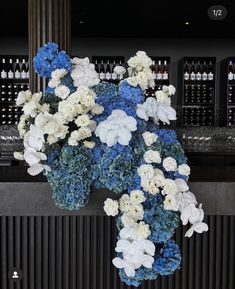 a bunch of blue and white flowers in a vase on a counter top with wine bottles behind it