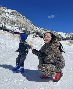 a woman kneeling down next to a small child on top of snow covered ground with mountains in the background