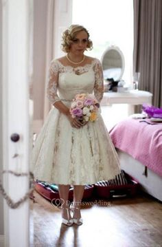 a woman in a white dress is standing near a bed and looking at the camera