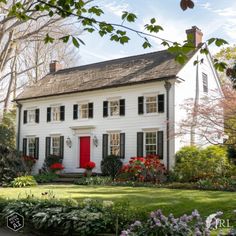 a white house with black shutters and red door in the front yard surrounded by flowers